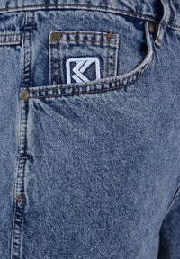 Denim jeans with a light blue wash, featuring a small white logo patch on the back pocket and copper-colored hardware details.