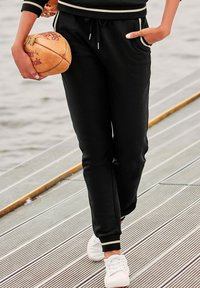 Schwarze Sweatpants mit Seitentaschen, elastischem Bund und Zugband, kombiniert mit weißen Turnschuhen. Die Hose hat weiße gestreifte Akzente.