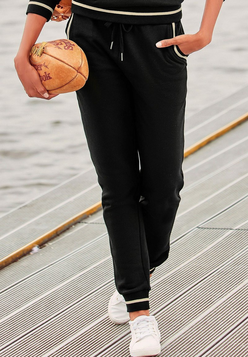 Schwarze Sweatpants mit Seitentaschen, elastischem Bund und Zugband, kombiniert mit weißen Turnschuhen. Die Hose hat weiße gestreifte Akzente.