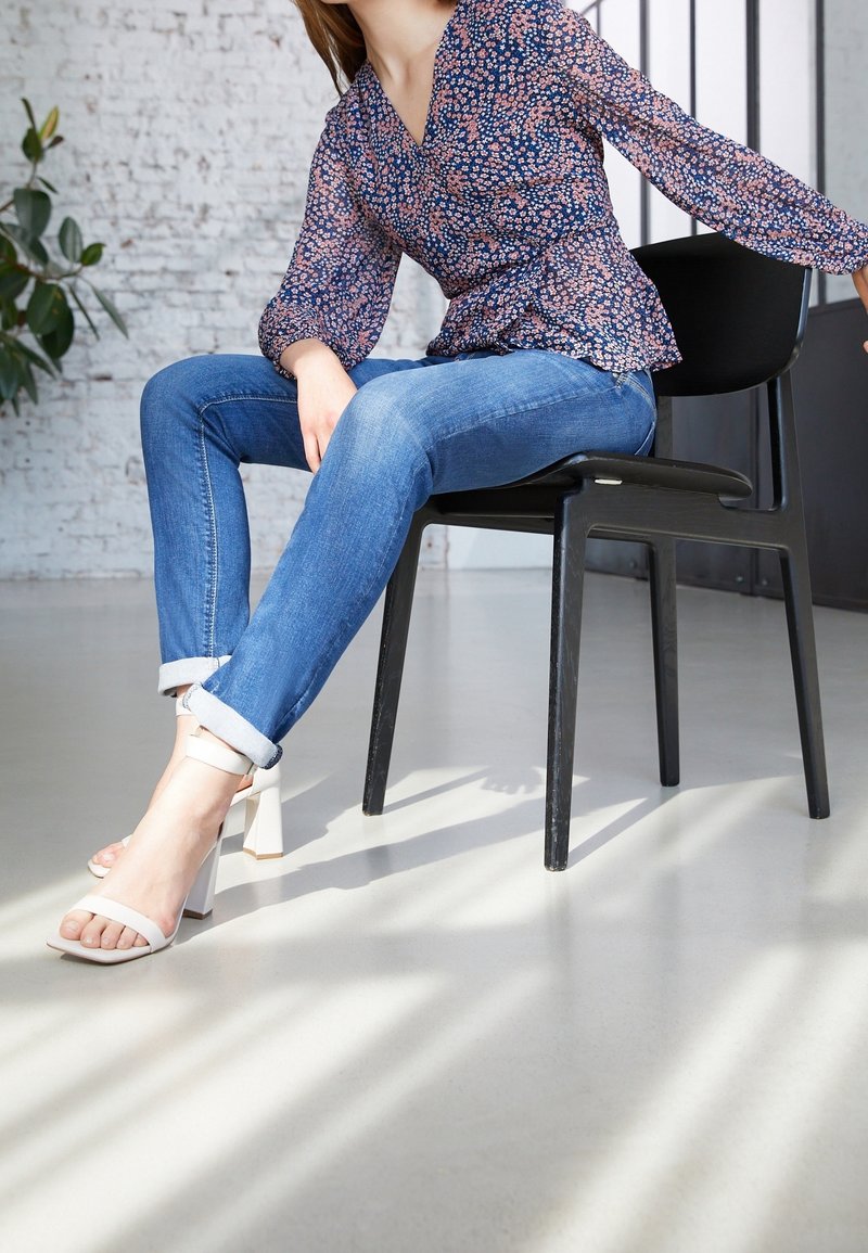 Bluse mit floralem Muster in Marineblau und langen Ärmeln; enganliegende blaue Jeans; weiße Sandalen mit Blockabsatz; sitzt auf einem schwarzen Stuhl in einem gut beleuchteten Raum.