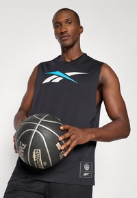 Μαύρη αμάνικη μπλούζα μπάσκετ με μπλε και λευκό λογότυπο, υφασμάτινη υφή και ετικέτα στο πλάι. Το μοντέλο κρατάει μια μαύρη μπάλα μπάσκετ με σχέδια για καλύτερη πρόσφυση.