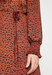 Robe couleur rouille avec imprimé léopard noir et beige, manches longues froncées avec poignets à volants, et nœud de ceinture mince sur le côté.