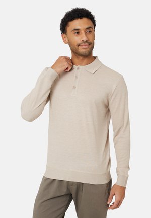 Man draagt een beige polo met lange mouwen en een grijze broek, die zachtjes de kraag aanraakt met een neutrale achtergrond.