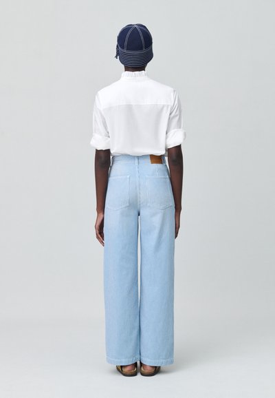 Chemise blanche à manches retroussées, associée à un pantalon taille haute rayé bleu clair. Un chapeau bleu foncé avec des rayures blanches complète le look.