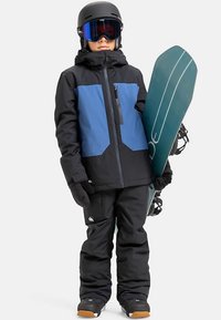 Sort og blå isoleret snowboardjakke med lynlås, kombineret med sorte bukser og handsker. Et snowboard holdes i højre hånd.