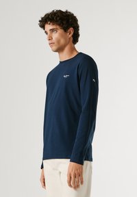 Ung mand med krøllet hår iført en mørkeblå langærmet skjorte og lysebeige bukser, der kigger til siden mod en ensfarvet baggrund.