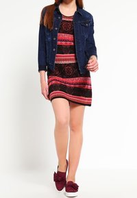 Veste en denim portée sur une robe à motif avec des designs rouges, roses et noirs. Associée à des chaussures à enfiler bordeaux. Textures lisses dans l'ensemble.