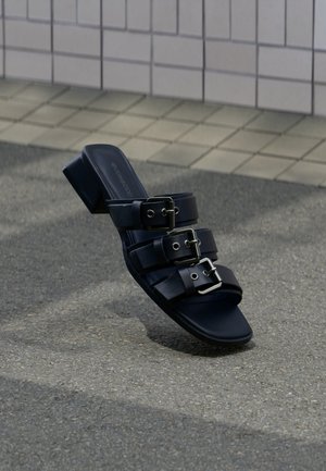 Μαύρο σανδάλι mule με ανοιχτά δάχτυλα, τρεις λουράκια με αγκράφες και χαμηλό τακούνι, τοποθετημένο σε επιφάνεια από άσφαλτο κοντά σε πλακόστρωτο πεζοδρόμιο.