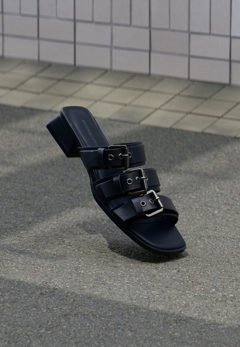 Μαύρο σανδάλι mule με ανοιχτά δάχτυλα, τρεις λουράκια με αγκράφες και χαμηλό τακούνι, τοποθετημένο σε επιφάνεια από άσφαλτο κοντά σε πλακόστρωτο πεζοδρόμιο.