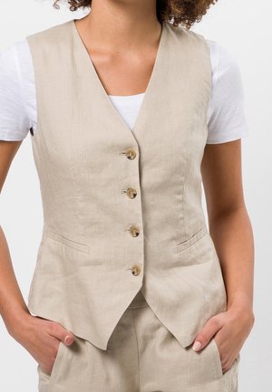 Frau trägt beige Knopfweste über weißem Kurzarmhemd mit den Händen in den Taschen der passenden beigen Hose, gezeigt vom Hals bis zur Hüfte.