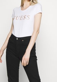 Hvid kortærmet t-shirt med "GUESS" trykt i roseguld glimmer over brystet, parret med sorte højtaljede jeans.