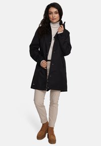 Juodas nepralaidus vandeniui paltas su užsegimais, aukšta apykakle ir šoninėmis kišenėmis, derinamas su smėlio spalvos kelnėmis ir rusvų raištelių aukštakulniais batais. Lygus paviršius.