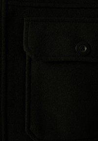 Černá vlněná látka s jemnou texturou, obsahující plochou kapsu a kulatý černý knoflík. Čistý design s jemnými detaily šití.