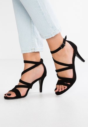 Kvinde fødder iført sorte remsandaler med høj hæl, parret med lyseblå cropped jeans på et hvidt gulv.