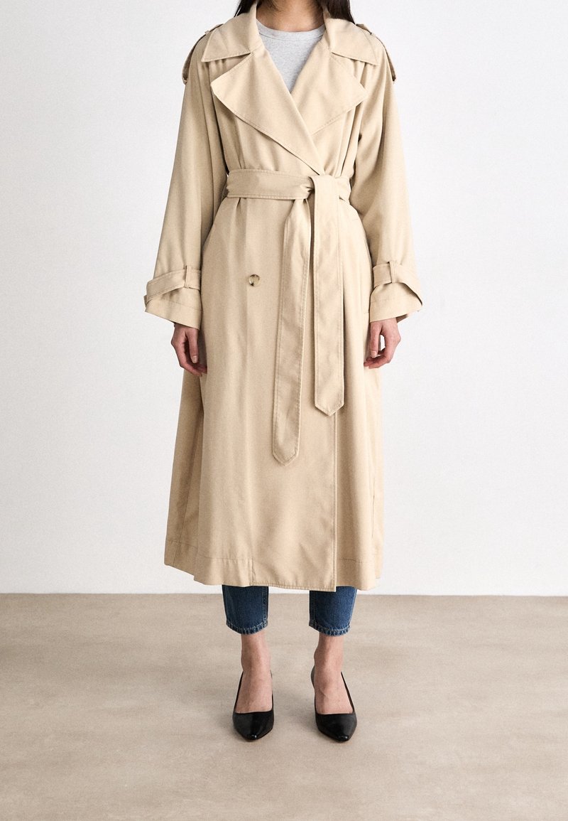 Femme portant un trench-coat long beige avec ceinture, associé à un jean bleu coupé au-dessus de la cheville et des escarpins noirs à bout pointu, debout sur un sol uni.