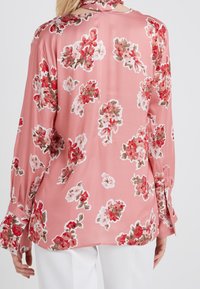 Blusa floreale rosa con maniche allungate e polsini con bottone, realizzata in tessuto liscio decorato con grandi motivi floreali rossi e bianchi.