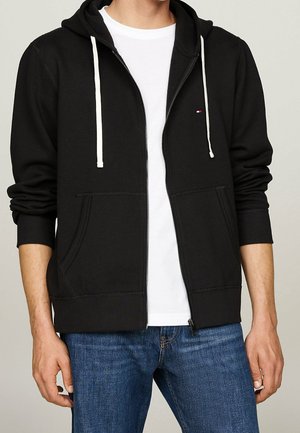Person iført sort lynlåstrøje med hvide snore over en hvid t-shirt og blå jeans, stående mod en enkel baggrund.