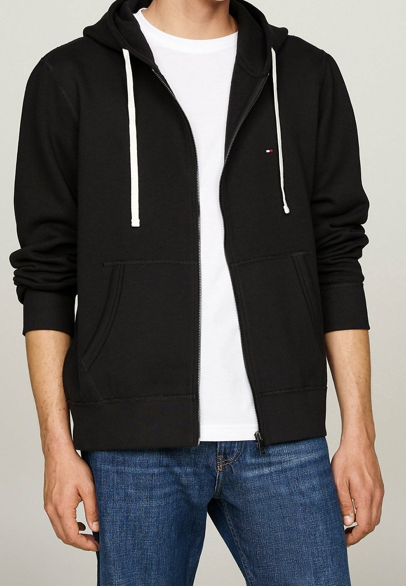 Personne portant un sweat à capuche noir à fermeture éclair avec des cordons blancs, un t-shirt blanc et un jean bleu, debout devant un fond uni.