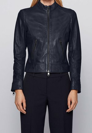 Femme portant une veste en cuir noire cintrée avec une fermeture éclair sur le devant et des poches zippées, associée à un pantalon noir ajusté.