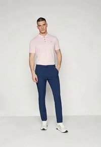 Roupa masculina com uma polo rosa clara com colarinho e logótipo, combinada com calças slim-fit azul-marinho e ténis brancos.