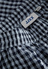 Tissu à carreaux bleu marine et bleu clair avec taille élastique ; l'étiquette indique "JJXX," fabriqué en Inde. Texture douce, design décontracté.