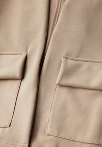 Beige mikina s měkkou texturou, která má dvě přední kapsy a hladký zip uprostřed.