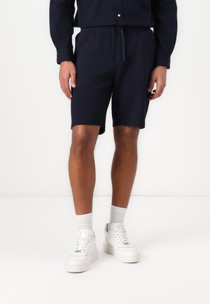 Marineblå bomuldsshorts med elastisk talje og snøre. Designet med en lige pasform, der slutter over knæet, parret med hvide sneakers.