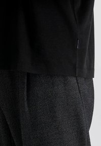 Schwarzes Baumwoll-T-Shirt mit kurzen Ärmeln, kombiniert mit strukturierter, dunkelgrauer Stoffhose, die ein kleines Etikett an der Seite hat.