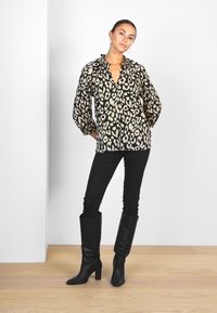 Blusa leopardata nera e beige con maniche lunghe, abbinata a jeans skinny neri e stivali neri alti fino al ginocchio su un pavimento di legno.