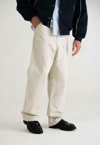 Pantaloni bej cu croială largă, cu cusături în contrast, buzunare cu elemente din alamă, asortați cu pantofi negri și o jachetă închisă, pe un fundal neutru.