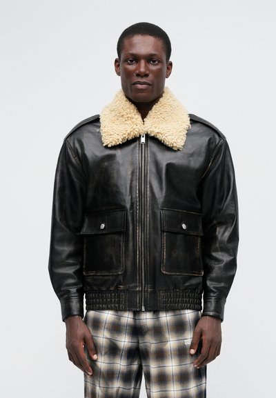 Veste en cuir noir avec un col en shearling beige, fermeture éclair à l'avant et deux poches poitrine ; finition texturée, ourlet côtelé. Portée avec un pantalon à carreaux.