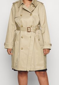Beige dubbelknäppt trenchcoat med midjeband, knäppta ärmslut och ficklock, bärs av en person mot en enkel bakgrund.