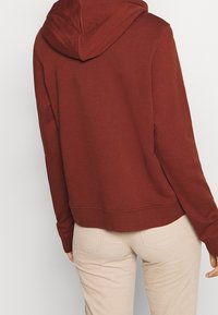 Maroon hoodie with a large hood, ribbed cuffs, and hem. Soft cotton blend fabric. Worn over light beige pants, showing a relaxed fit.