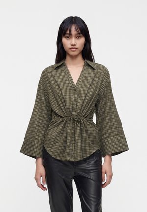 Femme portant une chemise à carreaux vert olive boutonnée, avec des manches larges et une taille avec cordon de serrage, associée à un pantalon en cuir noir.