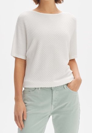 Witte, gestructureerde gebreide top met korte mouwen en een ronde hals. Gecombineerd met lichtgroene, strakke broek, handen in de zakken.