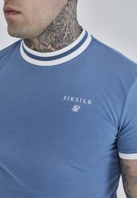 Kék rövidujjú póló fehér és sötétkék csíkozott bordával, apró fehér logóval a mellrészen, sima, puha textúrával.