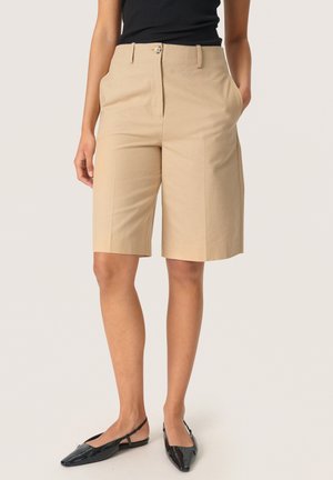 Kvinde iført beige knælange, skræddersyede shorts, sort ærmeløs top og sorte spidse slingback-flade sko, stående med hænderne i lommerne.