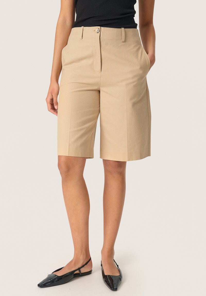 Femme portant un short beige sur mesure mi-cuisses, un haut noir sans manches et des ballerines slingback noires pointues, debout, les mains dans les poches.
