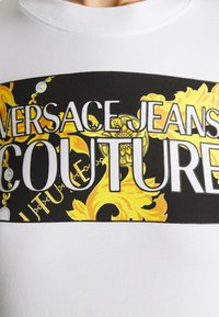 Vit bomullströja med en grafisk band i svart och guld. Inkluderar texten "VERSACE JEANS COUTURE" i fetstil och utsmyckade designelement.