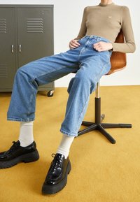 Haut à manches longues en maille côtelée couleur marron clair, jean bleu taille haute, chaussures noires épaisses, chaussettes crème. En arrière-plan, un casier vert et un sol jaune.