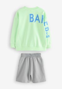 Lichtgroene sweatshirt met "BAKED" in het blauw gedrukt op de achterkant en mouwen, gecombineerd met grijze shorts met een elastische tailleband.