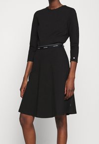 Černé šaty s dlouhými rukávy, kulatým výstřihem a rozšířenou sukní. Obsahují opasek s logem Calvin Klein a malé logové akcenty na rukávu.