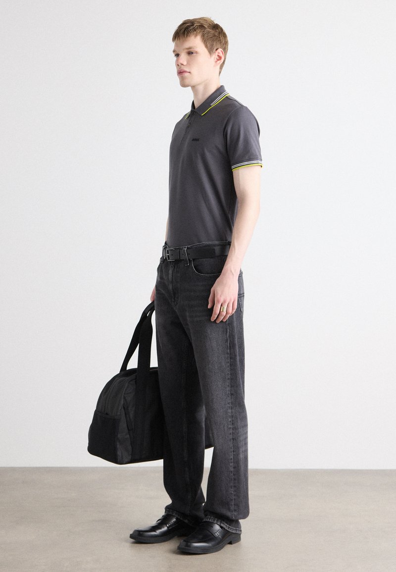 Kohlefarbendes Polohemd mit gelben und grau Akzenten, kombiniert mit dunklen Jeans und schwarzen Schuhen. Modell hält eine schwarze Sporttasche und steht auf einem neutralen Boden.