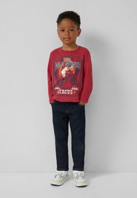 Junger Junge steht mit den Händen in den Taschen, trägt ein rotes Langarmshirt mit einem Tiger- und Motorradmotiv, dunkle Jeans und weiße Turnschuhe.