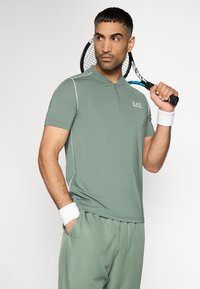 Πράσινο αθλητικό πόλο με φερμουάρ στον γιακά, κοντά μανίκια και λευκές λεπτομέρειες. Συνδυασμένο με αντίστοιχο παντελόνι, κρατώντας ρακέτα τένις.