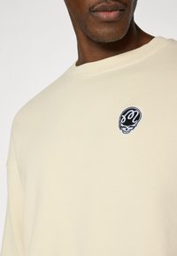 Ανοιχτό μπεζ φούτερ με λαιμόκοψη crew, με μικρό μαύρο λογότυπο κρανίου στο στήθος. Το ύφασμα έχει λεία υφή.