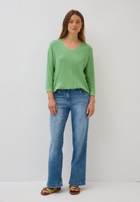 Zielona, długa bluzka z dekoltem w kształcie litery V w wykonaniu dzianiny, zestawiona z jasnoniebieskimi dżinsami w stylu dzwonów oraz karmelowymi sandałami o plecionej fakturze i detalach dekoracyjnych.