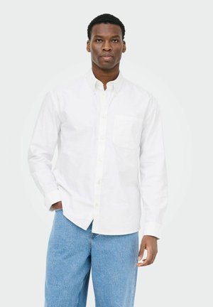 Mann trägt weißes Hemd mit Knöpfen und blaue Jeans, steht mit einer Hand in der Hosentasche vor einfarbigem Hintergrund.