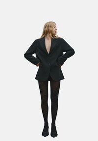 Czarny, oversize blezer z głębokim dekoltem w serek, stylizowany na krótkie czarne szorty i wzorzyste rajstopy z małymi srebrnymi kropeczkami, zestawione z czarnymi butami.