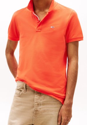 Homme portant un polo à manches courtes orange vif avec un petit logo sur la poitrine et un pantalon beige, debout avec une main dans la poche.